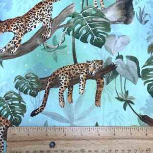 May include: Fabric with a jungle theme featuring leopards, lush green leaves, and branches against a light blue background. The leopards have brown and black spots. The ruler shows inches.