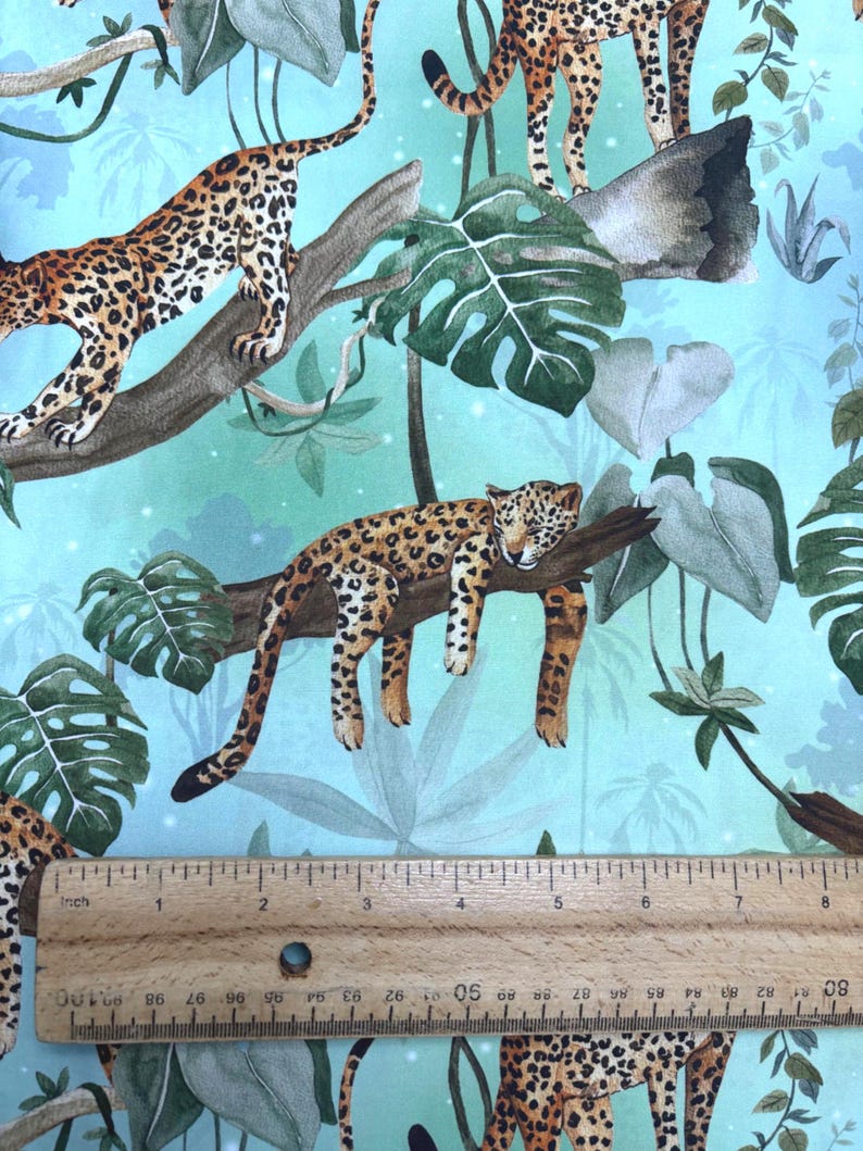 May include: Fabric with a jungle theme featuring leopards, lush green leaves, and branches against a light blue background. The leopards have brown and black spots. The ruler shows inches.