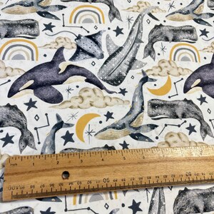 May include: A white fabric with a repeating pattern of whales, manta rays, stars, moons, and rainbows. The whales are gray and white, the manta rays are gray, the stars are black, the moons are yellow, and the rainbows are gray, yellow, and white.