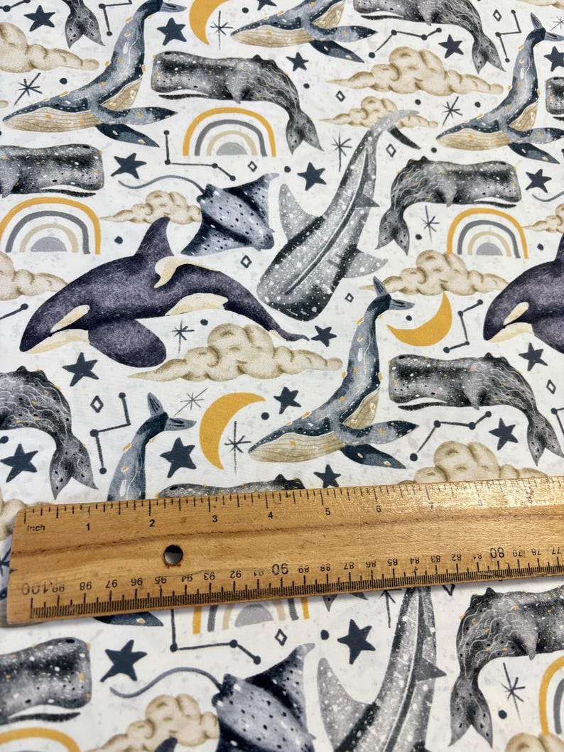 May include: A white fabric with a repeating pattern of whales, manta rays, stars, moons, and rainbows. The whales are gray and white, the manta rays are gray, the stars are black, the moons are yellow, and the rainbows are gray, yellow, and white.