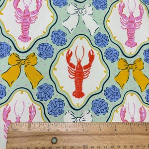 May include: Fabric with a repeating pattern of lobsters, bows, and seashells. The lobsters are pink and red, the bows are yellow and white, and the seashells are blue. The background is a light green color with small white dots. A wooden ruler is in the image.