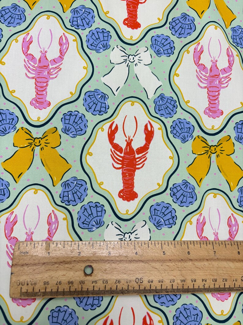 May include: Fabric with a repeating pattern of lobsters, bows, and seashells. The lobsters are pink and red, the bows are yellow and white, and the seashells are blue. The background is a light green color with small white dots. A wooden ruler is in the image.