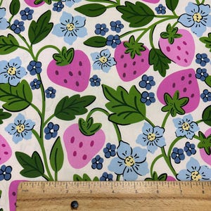 May include: Fabric with a repeating pattern of large pink strawberries, light blue flowers, and green leaves on a white background. A wooden ruler with inch markings is at the bottom of the image.