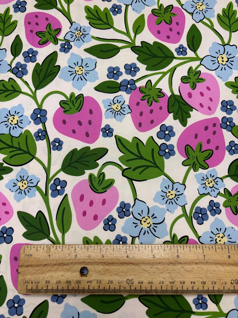May include: Fabric with a repeating pattern of large pink strawberries, light blue flowers, and green leaves on a white background. A wooden ruler with inch markings is at the bottom of the image.