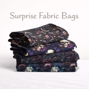 以下が含まれることがあります： 暗い花柄とスカル柄の生地バッグが4つ重ねられています。バッグは折りたたまれ、積み重ねられています。「Surprise Fabric Bags」の文字が上にあります。