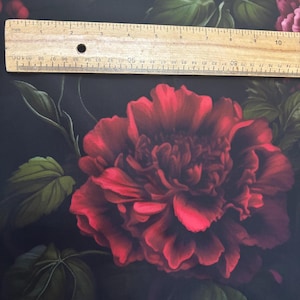 May include: A fabric featuring a large, deep red peony flower with dark green leaves on a black background, positioned beneath a wooden ruler that measures approximately 11 inches in length across the top of the floral pattern.