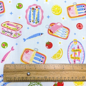 May include: Light blue fabric with a repeating pattern of sardines in cans, plates, and individual fish. The design includes lemons, tomatoes, olives, and flowers. A wooden ruler shows measurements in inches.