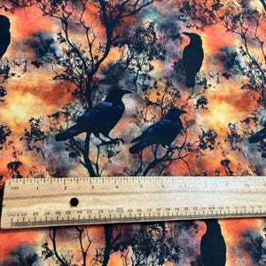 May include: A fabric with a repeating pattern of black crows silhouetted against a watercolor background of orange, yellow, and blue. The fabric has a subtle texture.