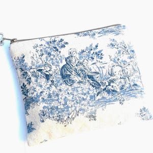 May include: A rectangular, off-white pouch with a blue toile print depicting a pastoral scene. The zipper is silver, and the pull is a star shape. The pouch features a classic toile pattern with figures and foliage.