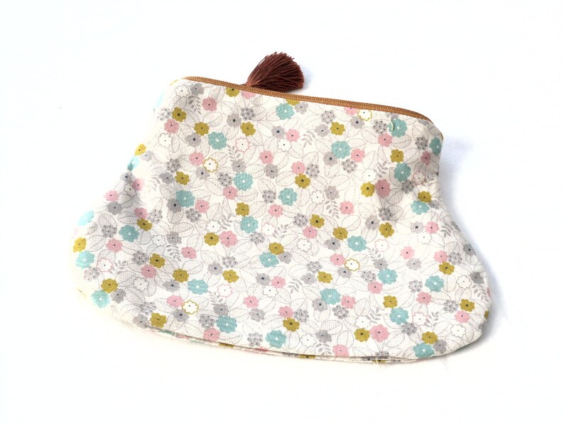May include: A white fabric pouch with a floral print in shades of pink, blue, and yellow. The pouch has a brown zipper and a brown tassel.