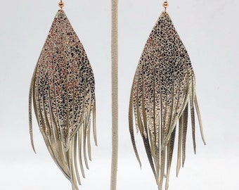 May include: A pair of gold leather fringe earrings with a textured, metallic finish. The earrings are teardrop shaped and have a long, flowing fringe.