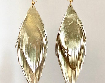 May include: Gold leather feather earrings with a layered design. The earrings are hanging from a wooden dowel.