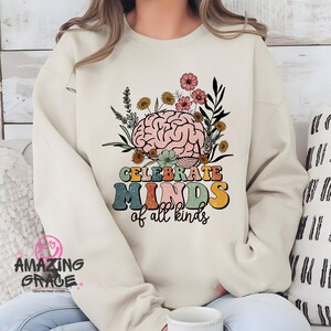May include: Cream-coloured sweatshirt with a colourful graphic. The design features a stylised brain surrounded by flowers and the text "CELEBRATE MINDS of all kinds" in a retro font. The sweatshirt has a crew neck and long sleeves.