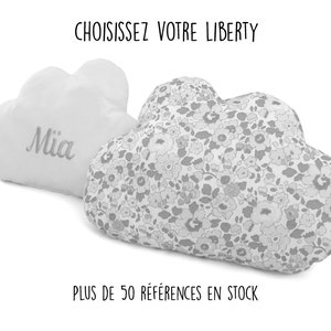 May include: Two white cloud-shaped cushions, one with the name "Mia" embroidered on it and the other with a floral pattern. The text "CHOISISSEZ VOTRE LIBERTY" is at the top of the image and the text "PLUS DE 50 RÉFÉRENCES EN STOCK" is at the bottom.