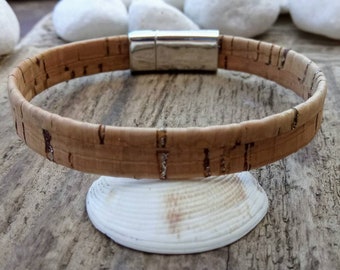 Puede incluir: Un brazalete de corcho marrón con un cierre magnético plateado. El brazalete está sobre una concha blanca.