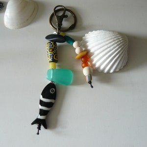Peut inclure: Un porte-clés artisanal avec un charm en forme de poisson rayé noir et blanc, des perles turquoise et jaunes, et un coquillage décoratif. Le porte-clés est attaché à un fermoir en métal de couleur bronze.