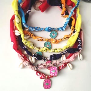 以下が含まれることがあります： カラフルな生地のネックレスのコレクション。各ネックレスは、赤、黄、青、ピンクなど、さまざまな色のねじれた生地で作られています。貝殻のアクセントと、目のデザインのターコイズペンダントが付いたネックレスもあります。