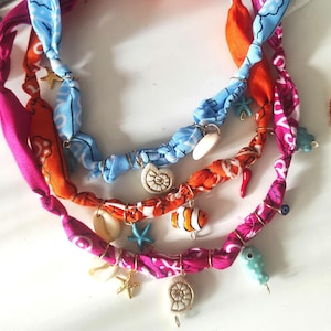 Peut inclure: Trois colliers en tissu colorés avec des breloques sur le thème de la mer. Les colliers présentent du tissu rose, orange et bleu avec des breloques en forme de coquillage, d'étoile de mer et de poisson. Les breloques sont fixées avec du fil doré.