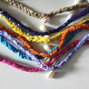 Peut inclure: Colliers en tissu tressé de différentes couleurs, dont rose, bleu, orange, jaune et violet. Chaque collier est orné d'un pendentif coquillage. Les colliers sont disposés sur une surface blanche.