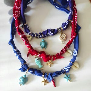 Könnte beinhalten: Drei handgefertigte Halsketten mit roten und blauen Paisley-Bandanas. Jede Halskette hat goldfarbene Drahtakzente und Anhänger, darunter Seesterne, Schildkröten und Muscheln. Die Halsketten haben ein nautisches Thema.