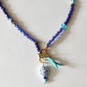 Könnte beinhalten: Eine Perlenkette aus dunkelblauen Saatperlen, durchsetzt mit kleinen goldfarbenen Zwischenelementen und hellblauen Akzenten. Die Kette verfügt über einen goldfarbenen Knebelverschluss mit einem weißen Keramikfisch-Anhänger und einem hellblauen korallenartigen Element.
