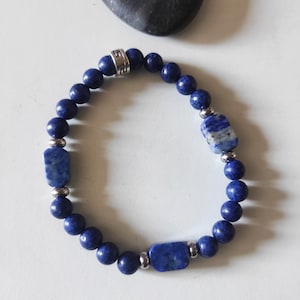Peut inclure: Un bracelet de perles bleu foncé avec des détails argentés. Le bracelet est composé de perles rondes et de pierres rectangulaires texturées. Une pierre sombre en forme de cœur est en arrière-plan.
