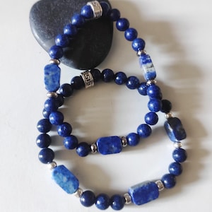 May include: Two beaded necklaces and a bracelet featuring deep blue lapis lazuli stones. The necklaces and bracelet have a mix of round and rectangular beads, with silver-coloured metal accents. The jewellery is displayed on a white surface.
