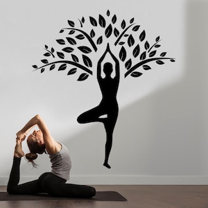 May include: A black silhouette of a person in a yoga tree pose is depicted on a white wall, with a tree design above. A person is doing yoga on a black mat. The image promotes wellness and fitness.