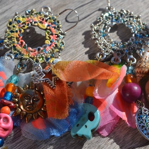 Può includere: Un paio di orecchini colorati con un design a filigrana argentata, che include una varietà di ciondoli, tra cui un sole, un teschio e una conchiglia. Gli orecchini sono adornati con perline, nastri e ritagli di tessuto in tonalità di rosa, arancio, blu e verde.