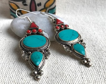 May include: Silver-coloured dangle earrings with turquoise and red accents. Each earring features an oval turquoise stone, a teardrop-shaped turquoise stone, and red coral-like beads. The earrings have an ornate design.