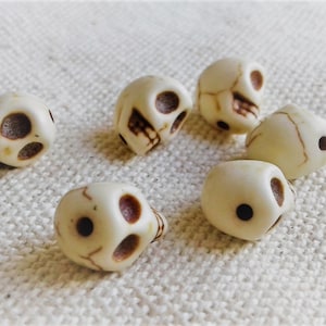 May include: Seven white skull beads with brown painted details. The beads are small and have a smooth, rounded surface.
