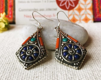 Può includere: Orecchini pendenti color argento con un design a forma di diamante. Gli orecchini presentano un motivo floreale blu centrale, dettagli arancioni e turchesi e un bordo argentato dettagliato. Sono esposti su una superficie bianca.