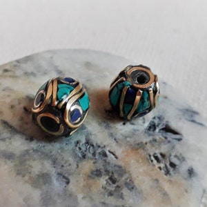 May include: Two round beads with intricate designs. The beads are made of brass and feature turquoise and blue accents.