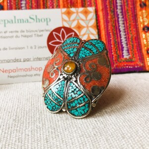 May include: A silver ring with a flower design. The flower is made of turquoise and coral stones. The ring is set on a white fabric background.
