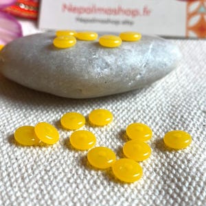 Puede incluir: Una colección de pequeñas cuentas redondas de vidrio amarillo dispersas sobre una superficie blanca texturizada. Algunas cuentas están dispuestas sobre una piedra gris lisa. La imagen también incluye una vista parcial de una tarjeta de visita con texto.