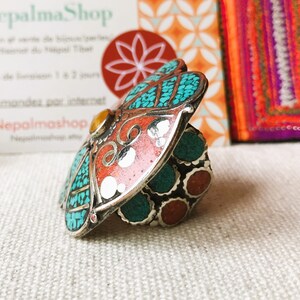 May include: A silver ring with a large, colorful, butterfly-shaped design. The butterfly has turquoise, red, and white accents. The ring is set on a white fabric background.