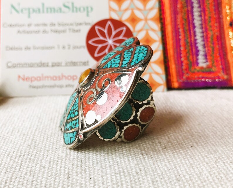 May include: A silver ring with a large, colorful, butterfly-shaped design. The butterfly has turquoise, red, and white accents. The ring is set on a white fabric background.