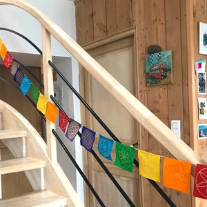 Peut inclure: Une guirlande de drapeaux carrés colorés suspendue le long d'une rampe d'escalier. Chaque drapeau est d'une couleur différente de l'arc-en-ciel, avec un motif géométrique doré. L'escalier est en bois clair et la rampe en métal foncé.