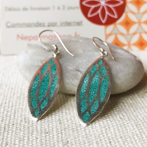 May include: Silver dangle earrings with a leaf-shaped design. The earrings feature a teal, glittery inlay within a silver frame. The earrings are displayed on a white surface.