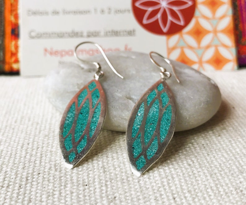 May include: Silver dangle earrings with a leaf-shaped design. The earrings feature a teal, glittery inlay within a silver frame. The earrings are displayed on a white surface.