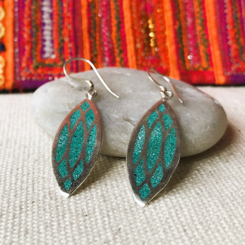 May include: A pair of silver earrings with a leaf-like design. The earrings feature a teal-colored, glittery inlay within a silver frame. The earrings are displayed on a white surface with a colorful textile in the background.