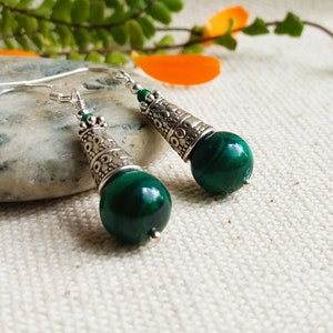 Può includere: Un paio di orecchini pendenti in argento con perline di pietra di malachite verde. Le perline sono rotonde e hanno una finitura liscia e lucida. Gli orecchini hanno un design a filigrana d'argento.