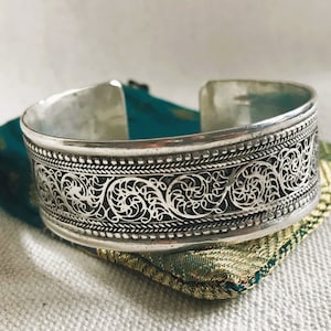 Peut inclure: Un bracelet argenté en forme de bangle avec un motif de filigrane complexe. Le bracelet est sur un fond de tissu vert et or.