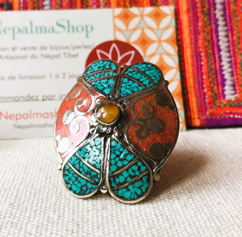 May include: A silver ring with a turquoise and coral inlay design. The ring is round and has a flower-like shape. The center of the ring has a small amber stone.
