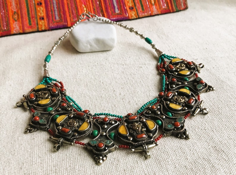 May include: A silver necklace with a multi-layered design featuring turquoise, red coral, and yellow stones. The necklace has a clasp closure.