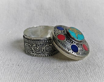 May include: A small, silver trinket box with an ornate design. The lid is decorated with turquoise, red, and blue stones.