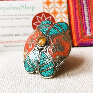 May include: A silver ring with a flower design. The flower is made of turquoise and coral stones. The ring is set on a white fabric background.