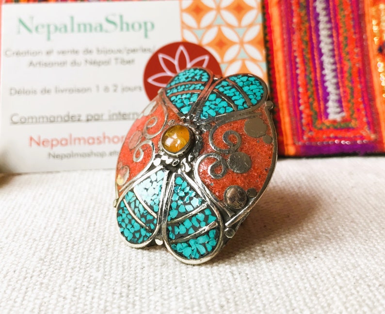 May include: A silver ring with a flower design. The flower is made of turquoise and coral stones. The ring is set on a white fabric background.