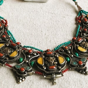 May include: A silver necklace with turquoise, red coral, and yellow stones. The necklace is made of intricate metalwork and has a beaded fringe.