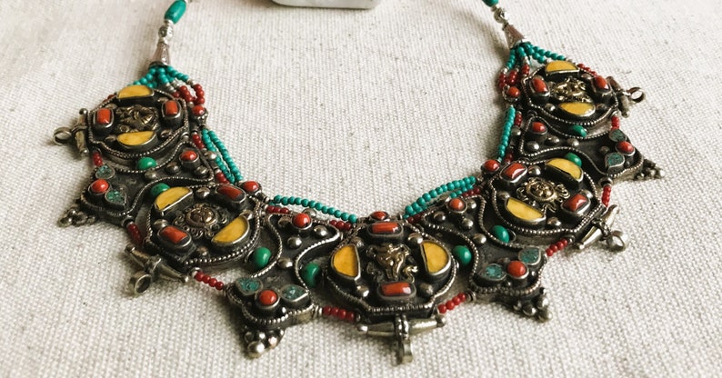 May include: A silver necklace with turquoise, red coral, and yellow stones. The necklace is made of intricate metalwork and has a beaded fringe.